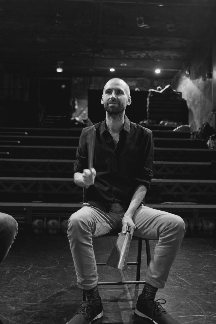 Lutz Woellert sitzt auf einer Theaterbühne und schlägt mit einem Stab auf eine Kuhglocke, die Hand in Bewegung